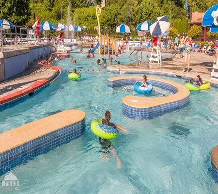 A bustling outdoor water park with multiple pools, a lazy river filled with people in inflatable tubes, colorful slides, splash features, lifeguard stands, and numerous visitors enjoying a sunny day.