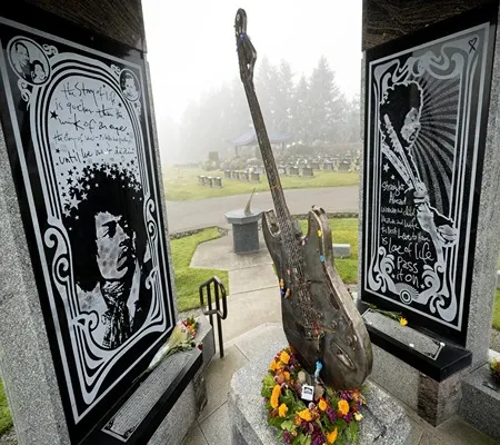 A memorial featuring etched portraits of Jimi Hendrix on black granite panels, flanking a bronze sculpture of a Stratocaster guitar adorned with flowers and guitar picks, situated in a cemetery with gravestones visible in the misty background.