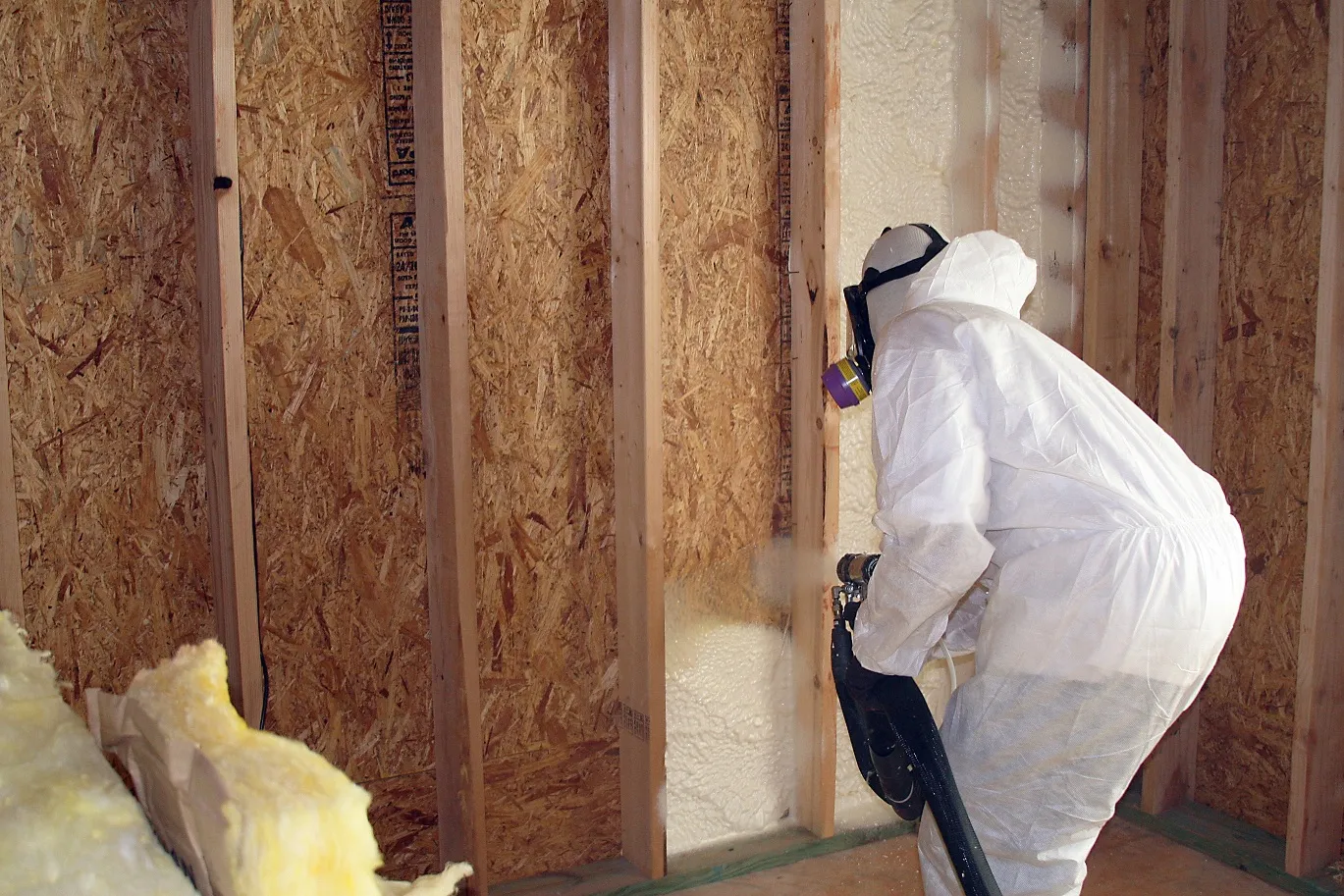 A person in a white protective suit, hood, and respirator is applying white spray foam insulation between the wooden studs of a wall in a building under construction. Some unfaced fiberglass insulation batts are visible on the floor.