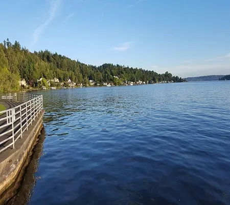 A calm, blue lake stretches out under a clear, light blue sky, bordered by a green, tree-lined shore with houses and a paved walkway with a white railing in the foreground.