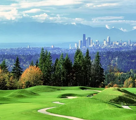 A lush green golf course with rolling hills and sand traps overlooks a distant cityscape of Seattle, framed by evergreen trees and a hazy mountain range under a partly cloudy sky.