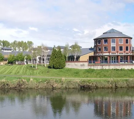 A modern community center building with a distinctive round brick tower is situated on a grassy bank overlooking a calm river, with trees lining the opposite shore under a partly cloudy sky.